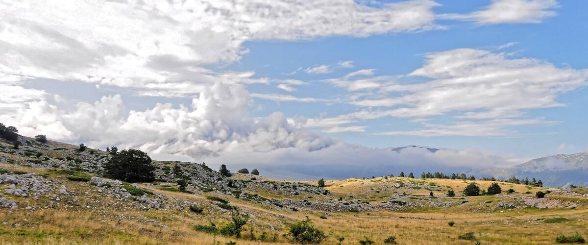 A mountainous landscape with rocky terrain, scattered trees, and a partly cloudy sky.