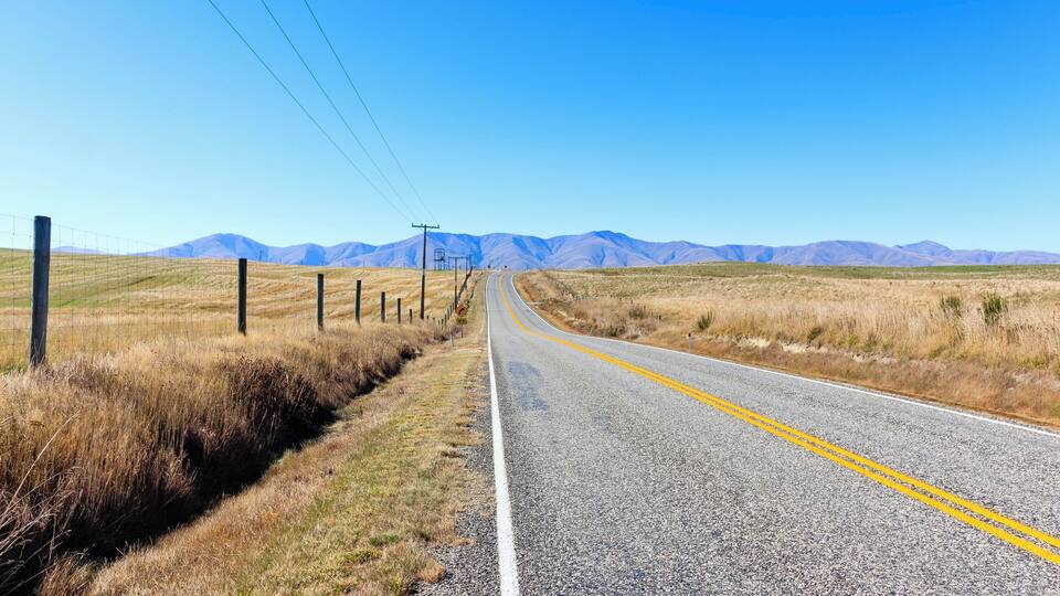 A two-lane road with a yellow center line, flanked by dry grass and a fence, leading towards distant mountains under a clear blue sky.