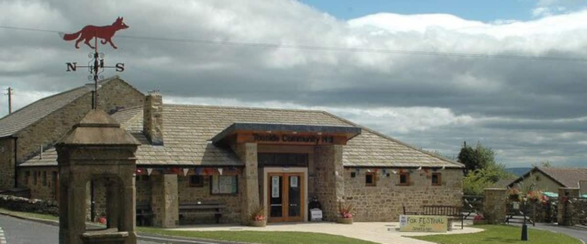 A stone building with a red fox on a weather vane, a sign for 'Tavistock Community Hall', and a 'For Sale' sign.