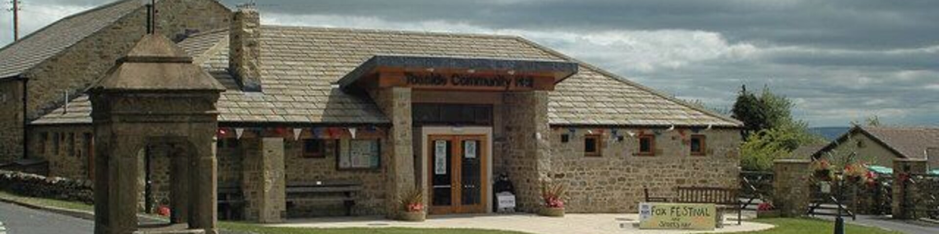 A stone building with a red fox on a weather vane, a sign for 'Tavistock Community Hall', and a 'For Sale' sign.