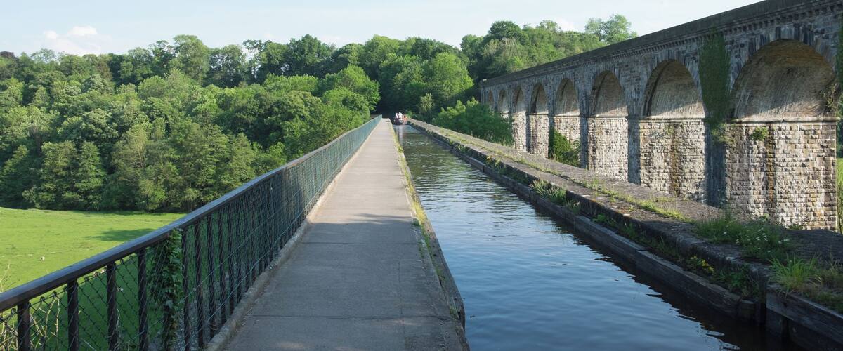 A stone bridge over a canal with a green fence on one side and trees in the background.