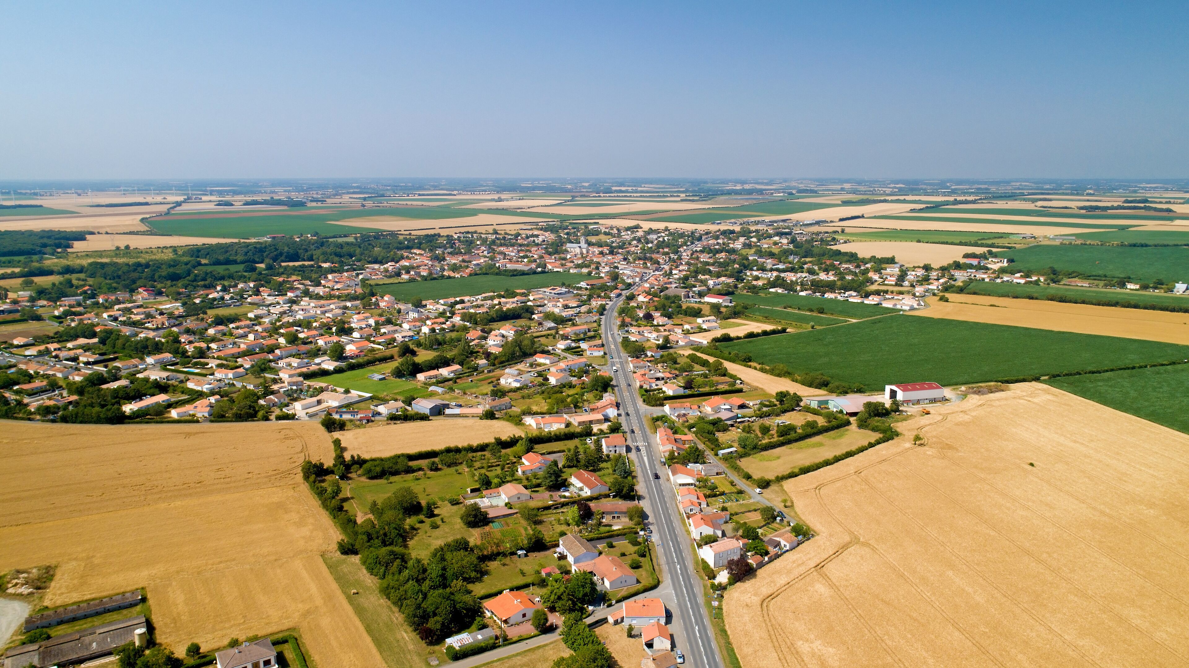 An aerial view of a rural town with a main road, fields, and wind turbines in the distance.