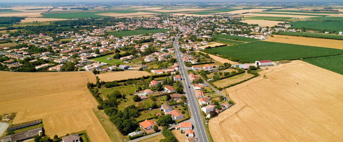 An aerial view of a rural town with a main road, fields, and wind turbines in the distance.