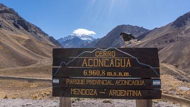 Um sinalizador com 'CERRO ACONCAGUA' e '6.960,8 m.s.n.m.' ao fundo de uma paisagem montanhosa.