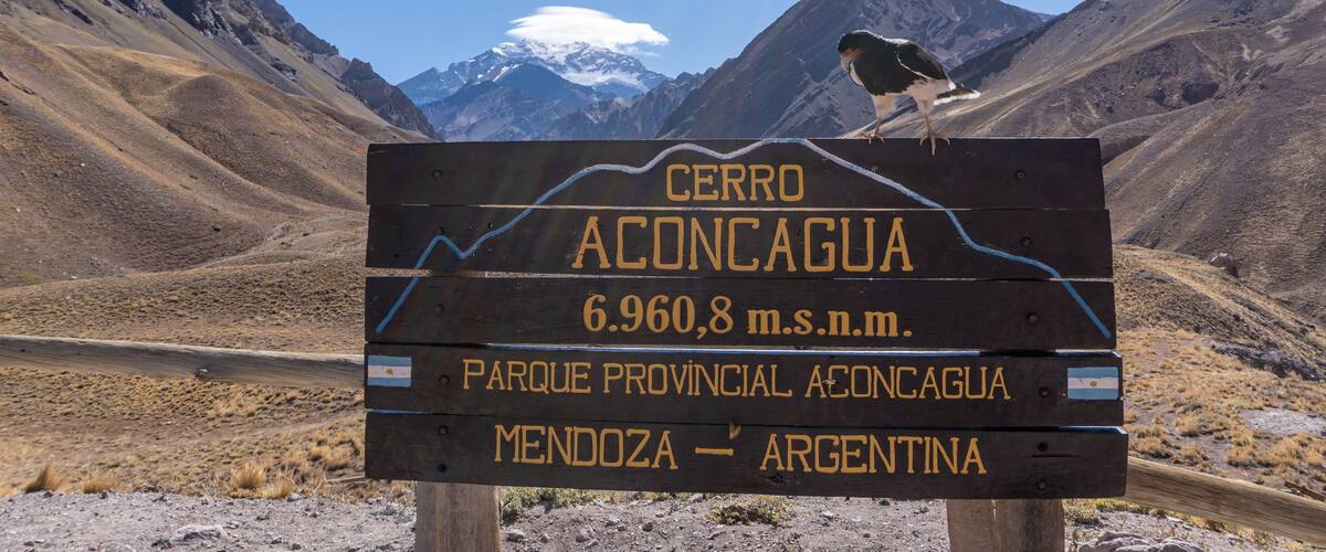 A signpost with 'CERRO ACONCAGUA' and '6.960,8 m.s.n.m.' in the background of a mountainous landscape.