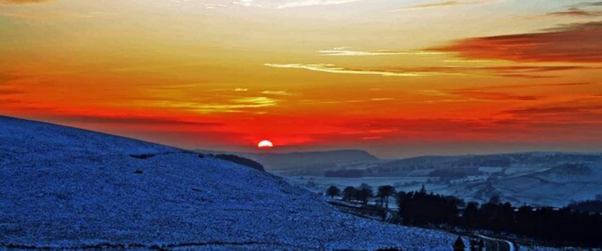 A sunset over a snow-covered landscape with a visible sun, distant hills, and a clear sky.