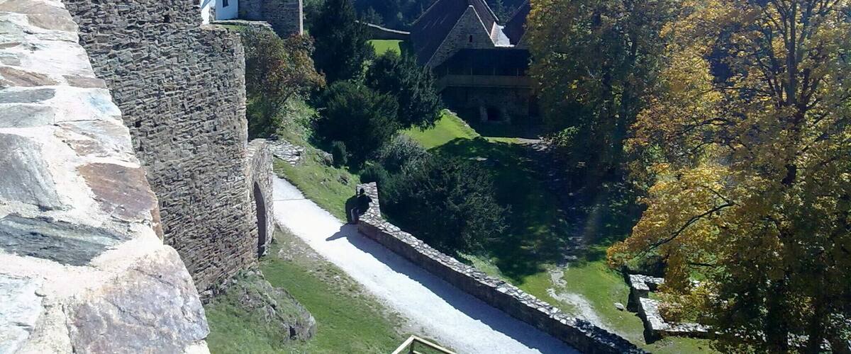 A stone castle with a white building adjacent to it, surrounded by a forested valley.