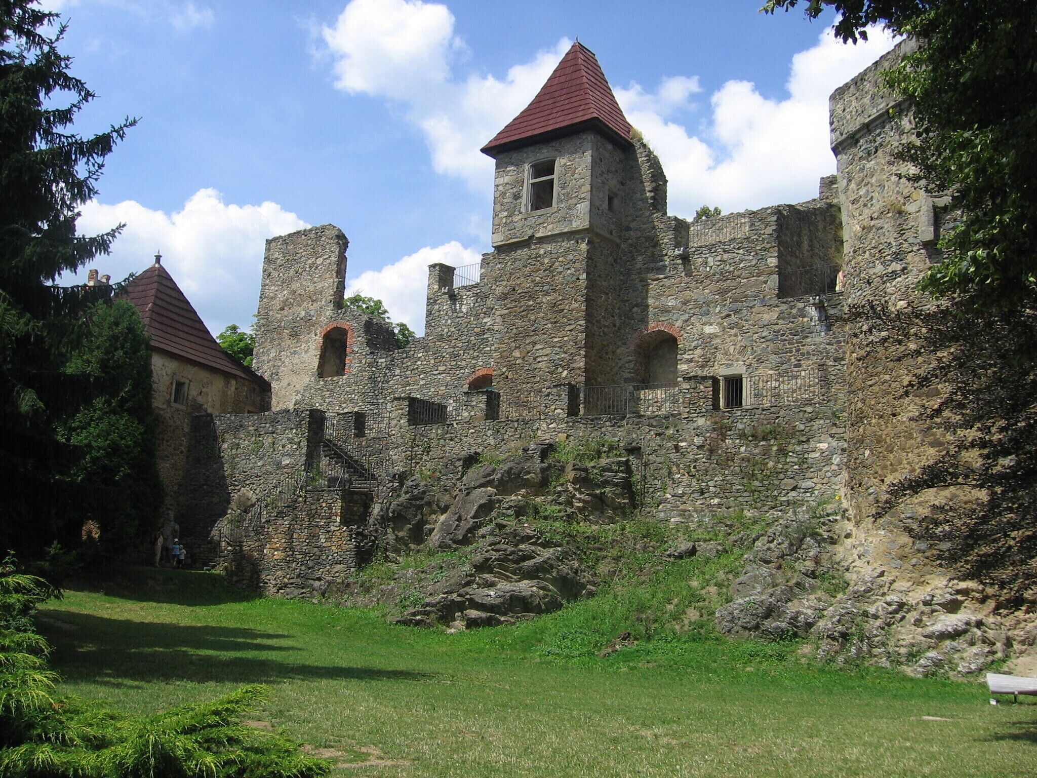 A historic stone castle with red-tiled roofs, surrounded by lush greenery and a clear blue sky.