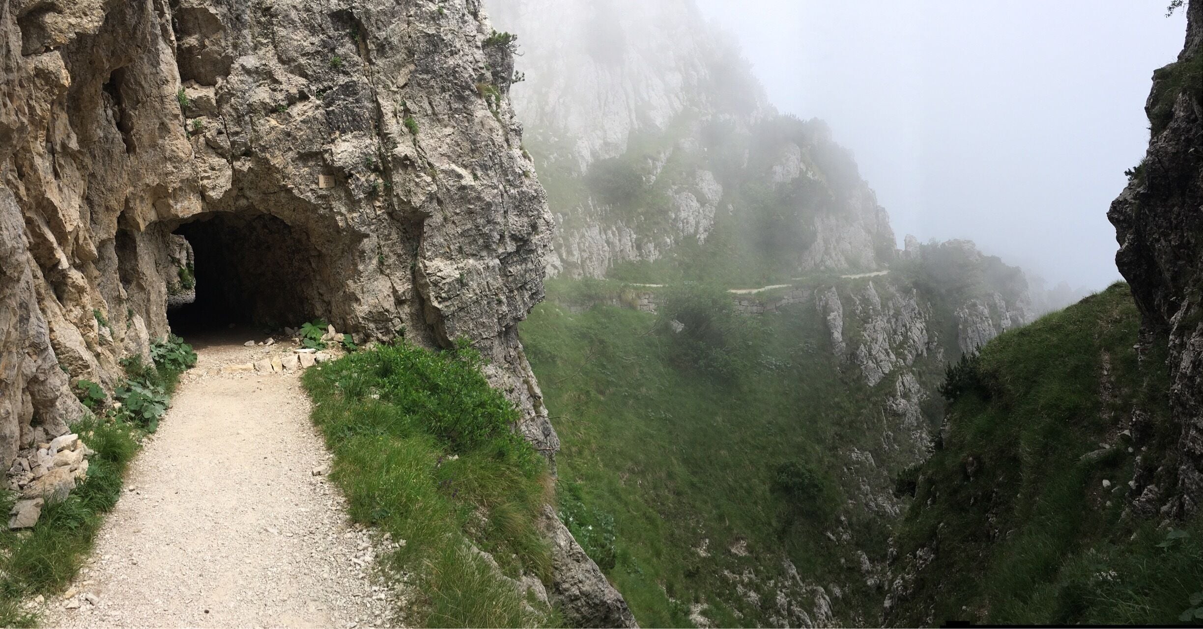 A rocky mountain path leading to a cave entrance surrounded by steep cliffs and lush greenery.