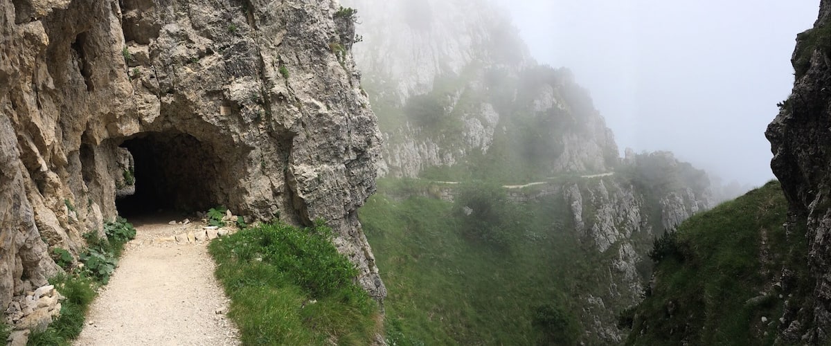 A rocky mountain path leading to a cave entrance surrounded by steep cliffs and lush greenery.