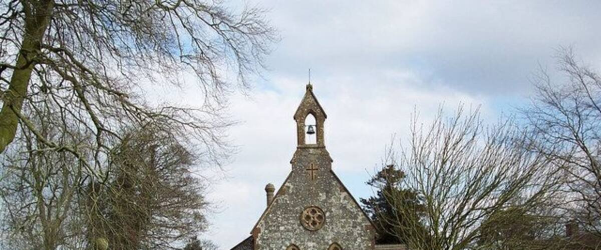 A stone church with a bell tower, surrounded by trees and a wooden fence.