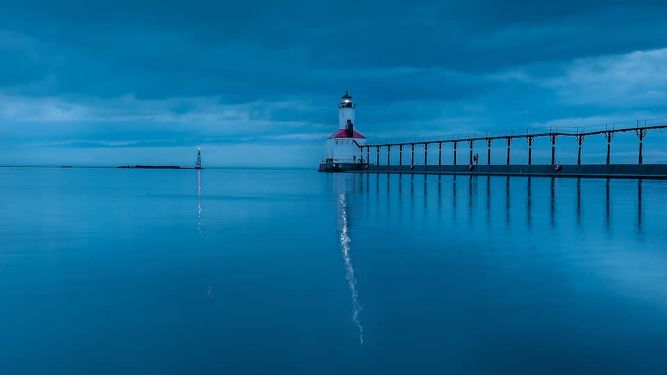 A lighthouse at the end of a pier with calm water reflecting the structure.