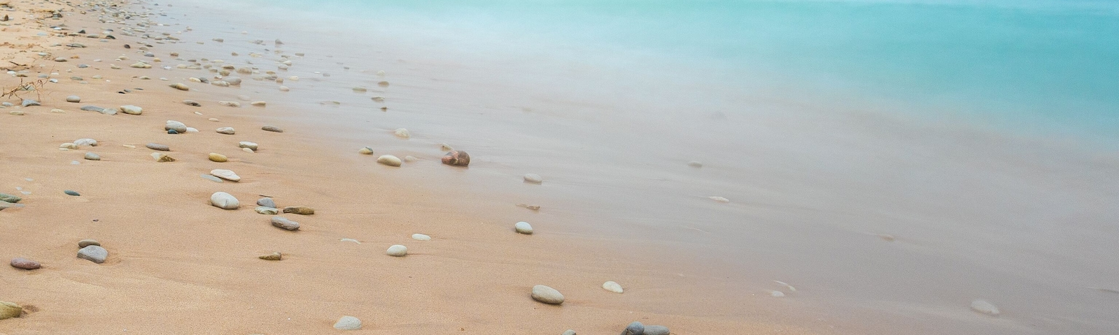 Uma praia arenosa com pedras espalhadas e águas turquesa calmas sob um céu claro.