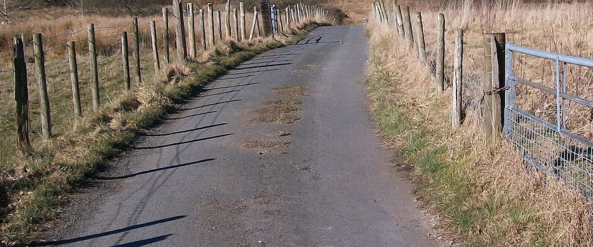 A narrow rural road flanked by wooden fences, leading through a hilly landscape with sparse vegetation and distant houses.