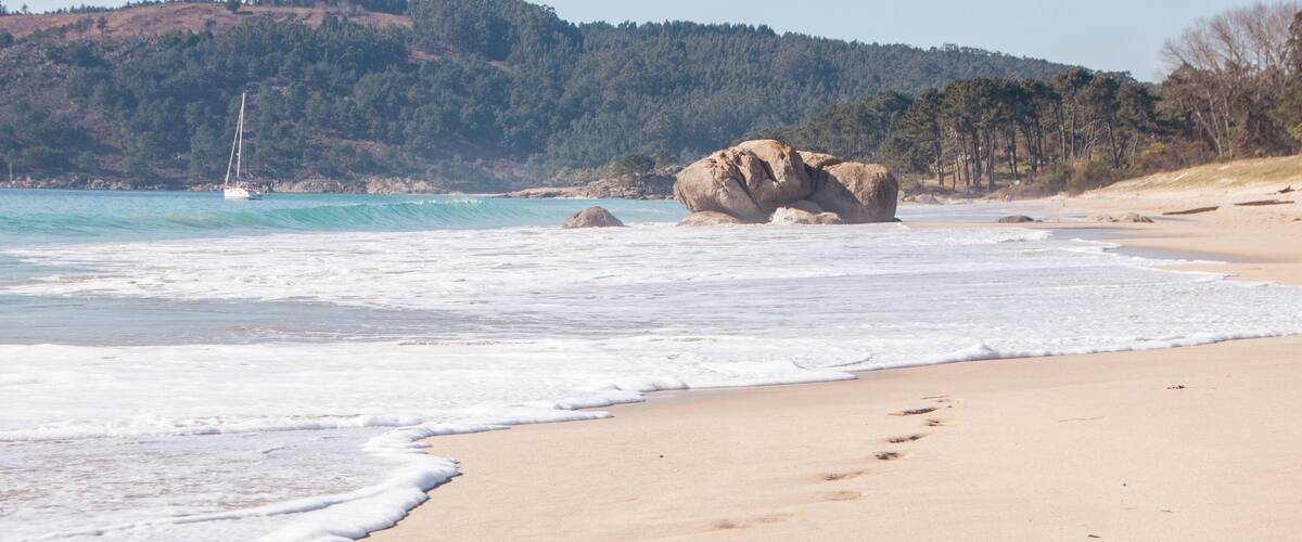 A sandy beach with footprints leading to a large rock formation, a sailboat in the water, and a forested hill in the background.