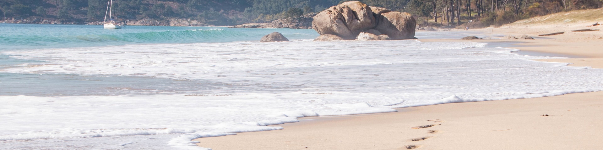 A sandy beach with footprints leading to a large rock formation, a sailboat in the water, and a forested hill in the background.