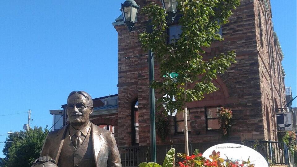 A bronze statue of a man sitting on a bench with a soccer ball, in front of a brick building with a clock tower.