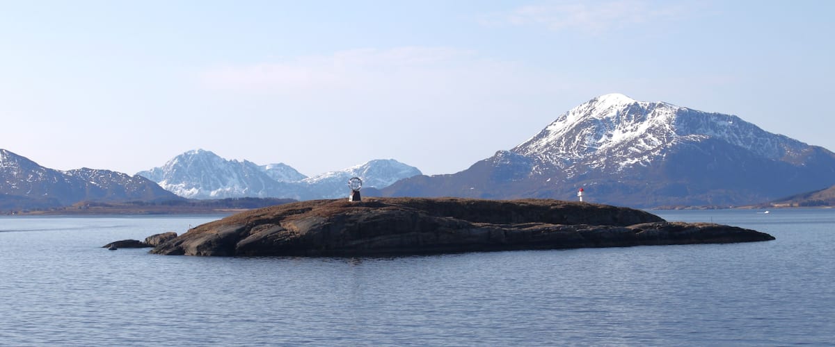 A rocky island with a lighthouse in the distance, surrounded by calm water and snow-capped mountains.