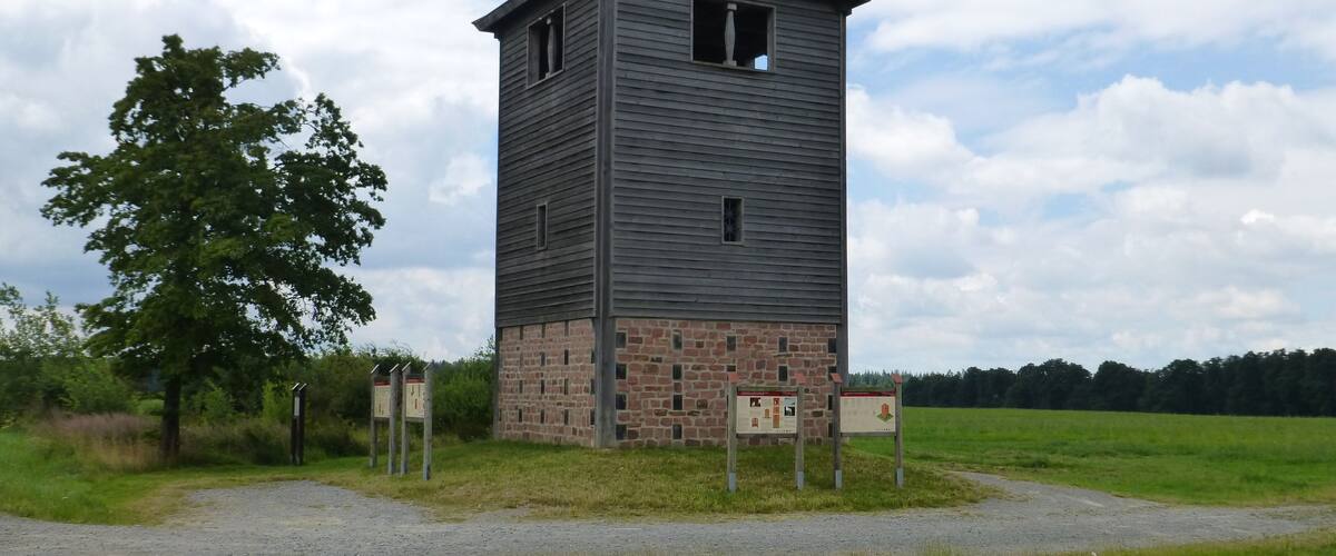A tall, narrow tower with a brick base and a wooden upper section, surrounded by a grassy field and trees.