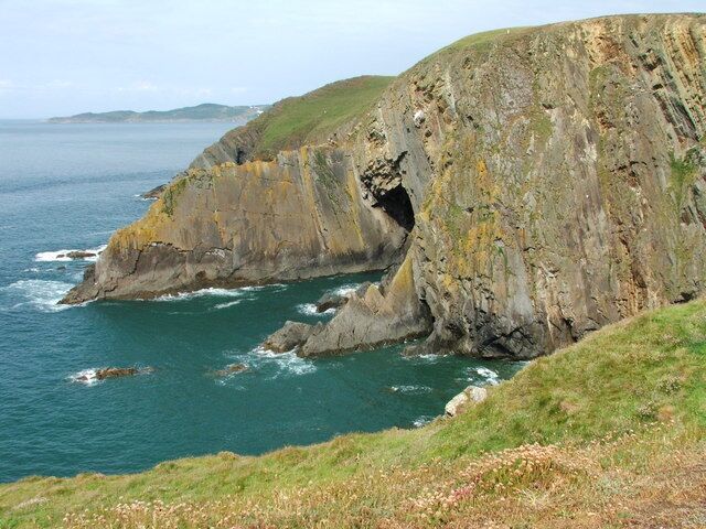 A coastal cliff with a cave entrance, grassy slopes, and the ocean.