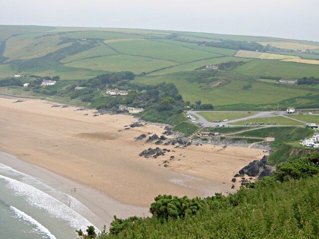 A coastal landscape with a sandy beach, rocky outcrops, and rolling green fields.