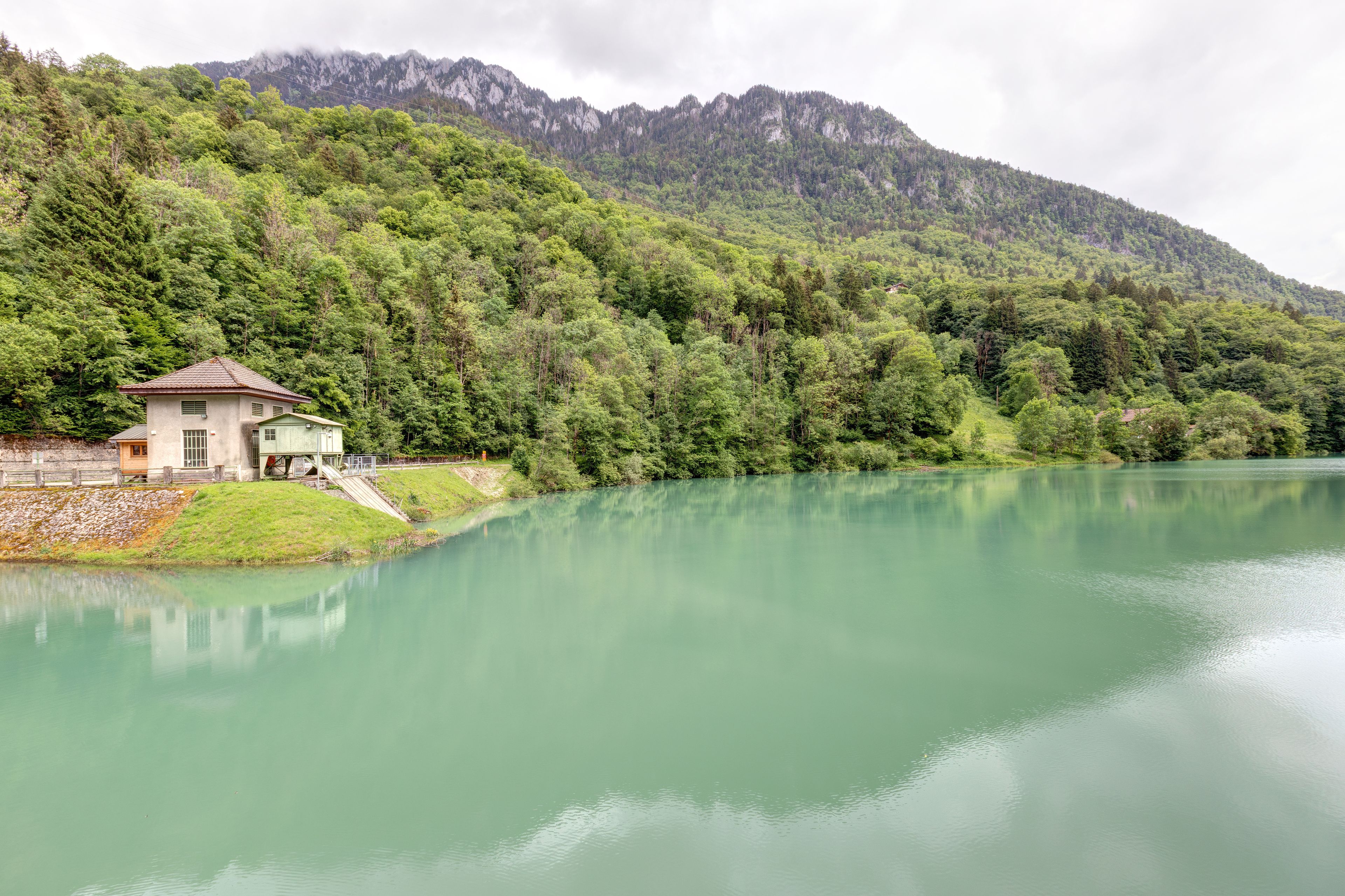 Een serene plas water met een klein huis aan de oever, omgeven door weelderige begroeiing en bergen op de achtergrond.