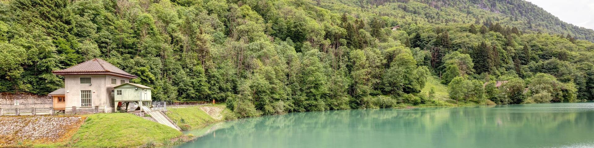 A serene lake with a small house on the shore, surrounded by lush greenery and mountains in the background.