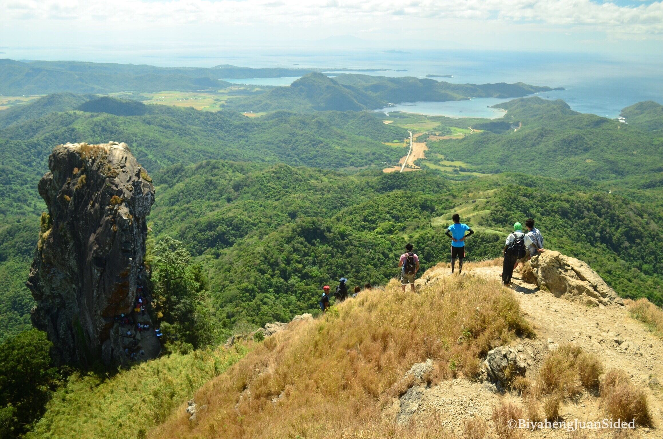 A mountainous landscape with hikers on a trail, a large rock formation, and a view of a coastal area in the distance.