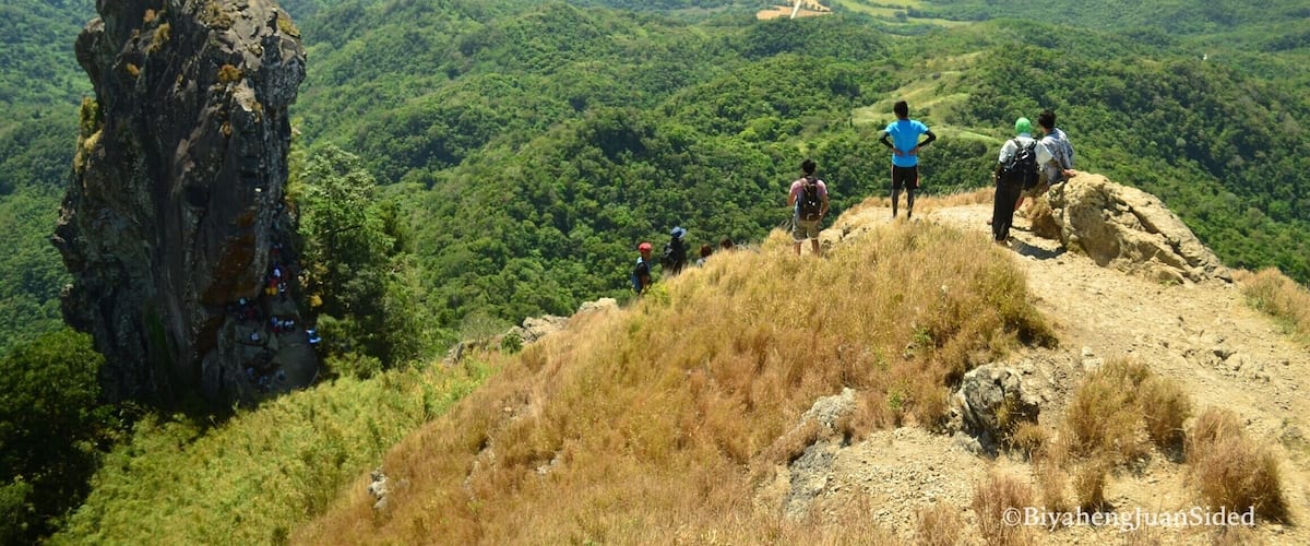 A mountainous landscape with hikers on a trail, a large rock formation, and a view of a coastal area in the distance.