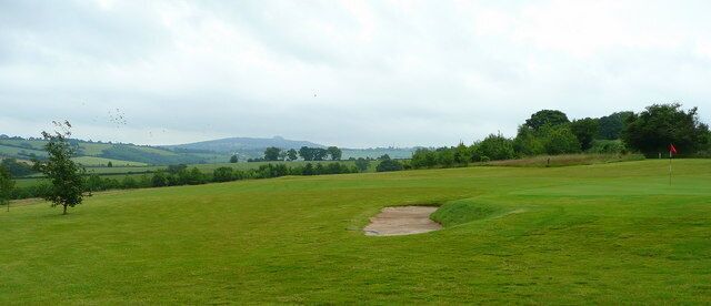 A golf course with a sand bunker, trees, and rolling hills in the background.