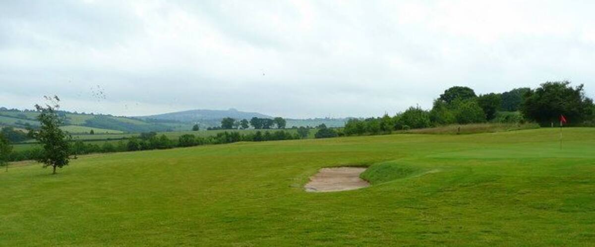 A golf course with a sand bunker, trees, and rolling hills in the background.