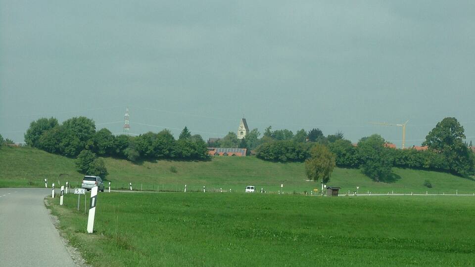 A rural road with a grassy field, a church, and wind turbines in the background.
