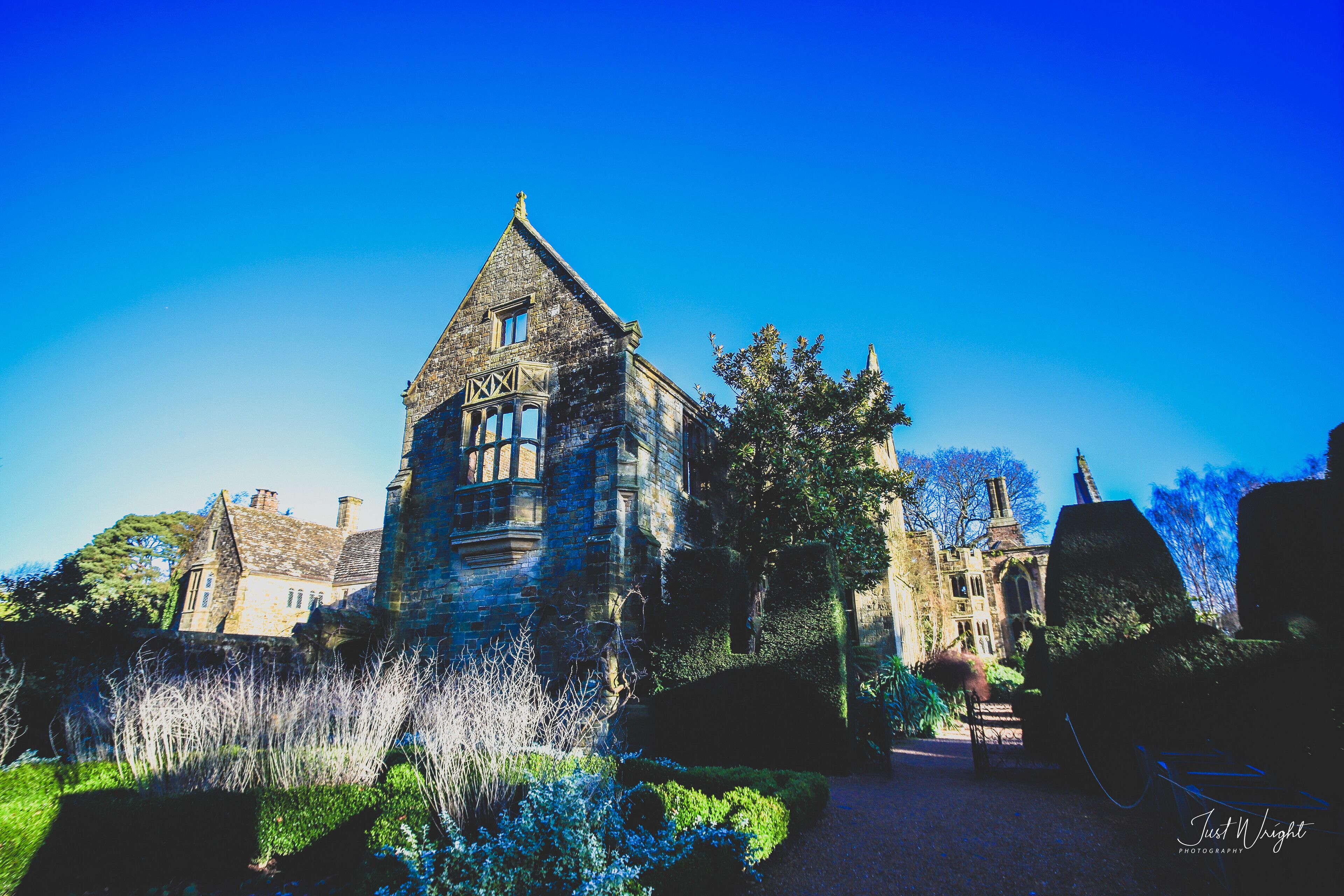 A historic stone building with a pointed roof and large windows, surrounded by well-maintained gardens and trees.