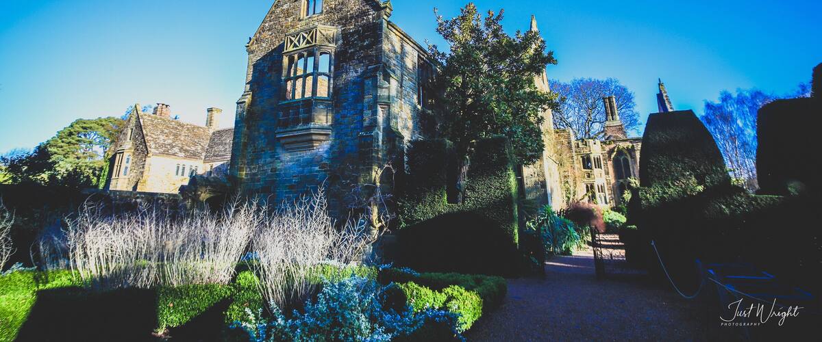 A historic stone building with a pointed roof and large windows, surrounded by well-maintained gardens and trees.