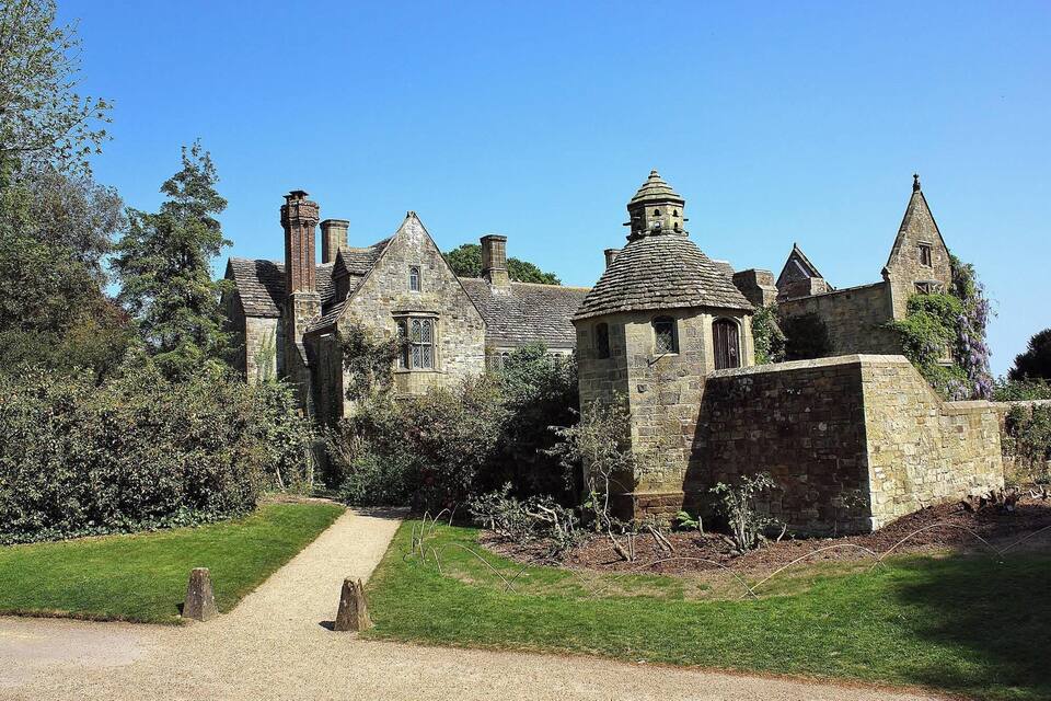 A historic stone building with multiple towers and a central dome, surrounded by lush greenery and a gravel path.