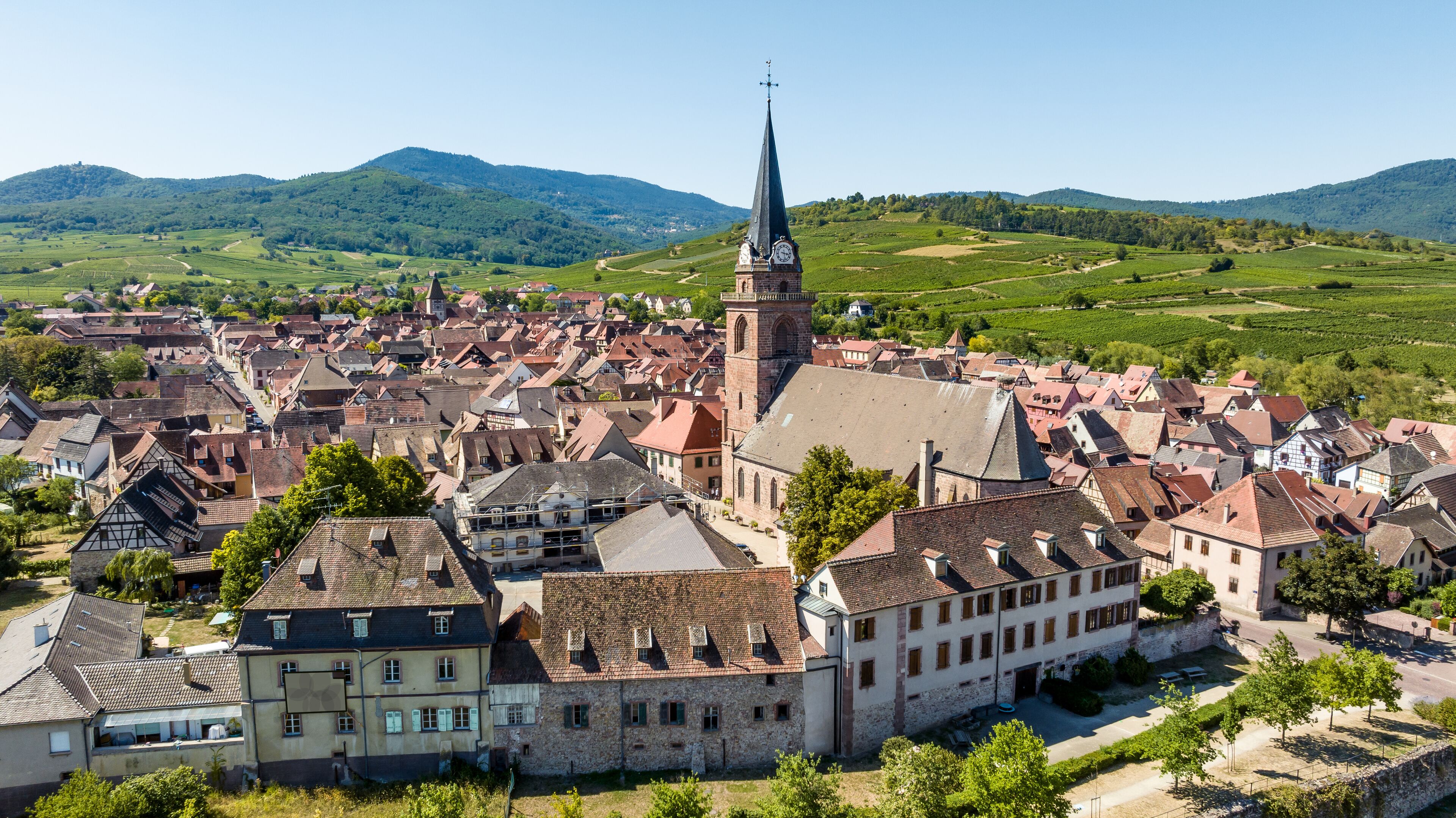 A historic town with a prominent church tower, surrounded by lush green hills and vineyards.