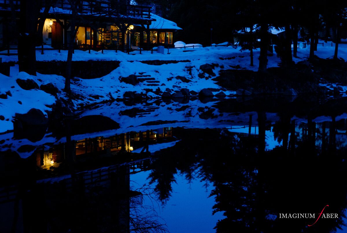 A snow-covered landscape with a lit cabin reflecting on a still body of water at night.