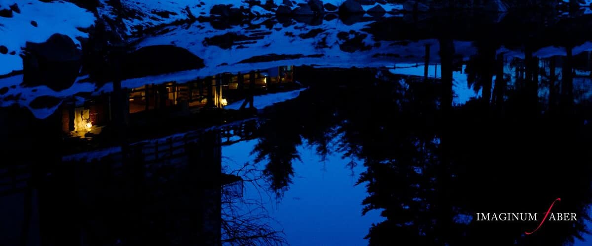 A snow-covered landscape with a lit cabin reflecting on a still body of water at night.