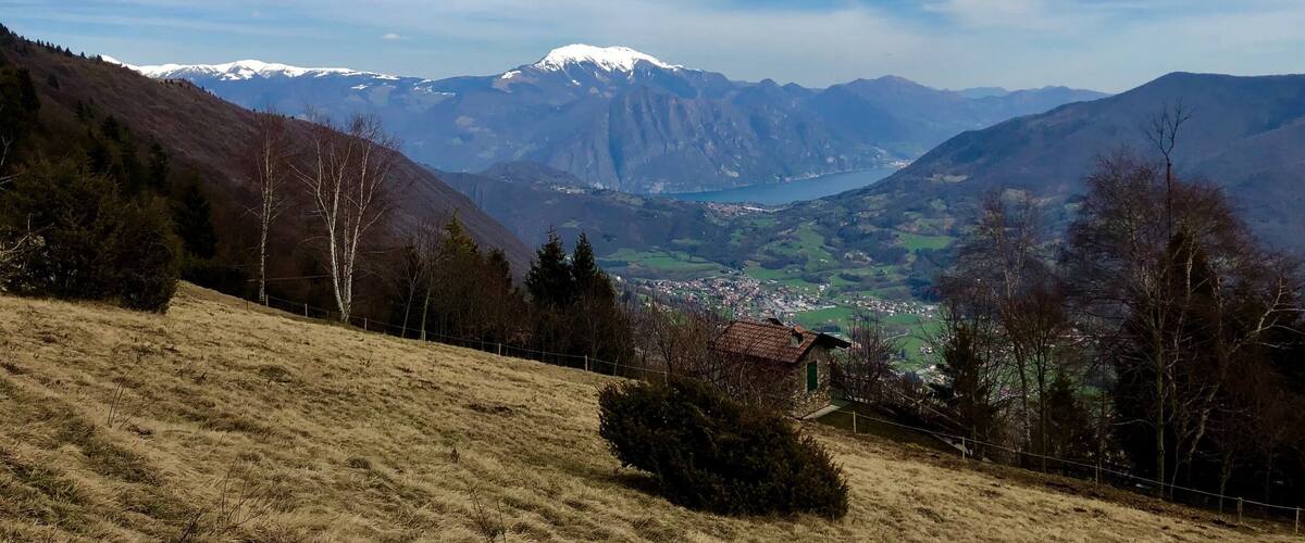 A mountainous landscape with a small house on a hill, a valley with a lake, and snow-capped mountains in the distance.