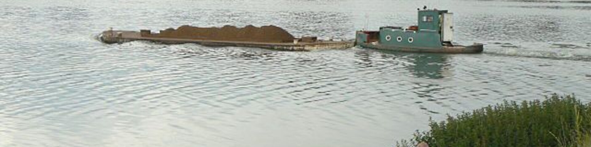 Uma barcaça em um rio, com alguém pescando na margem.