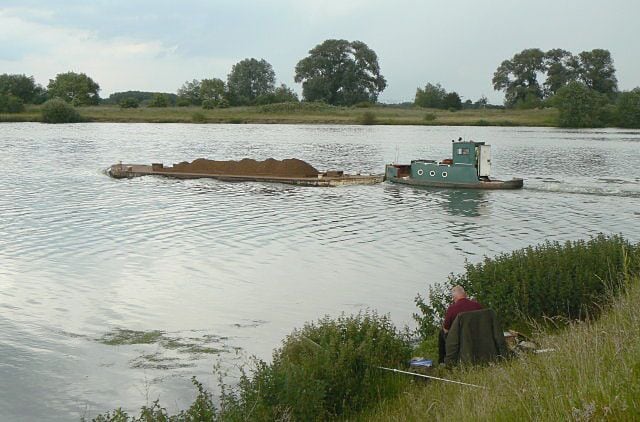 A barge on a river with a person fishing by the bank.