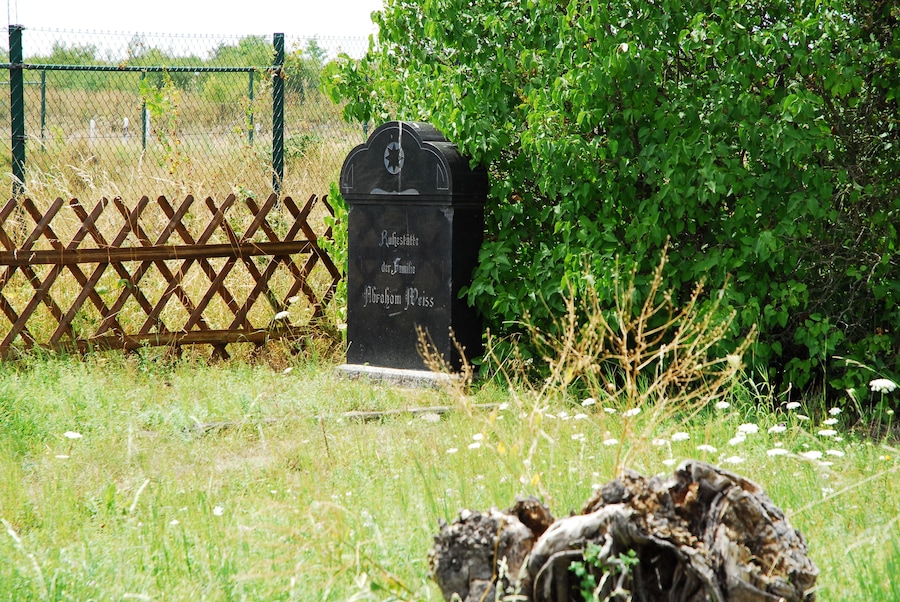 A headstone with inscriptions in a cemetery surrounded by greenery and a wooden fence.