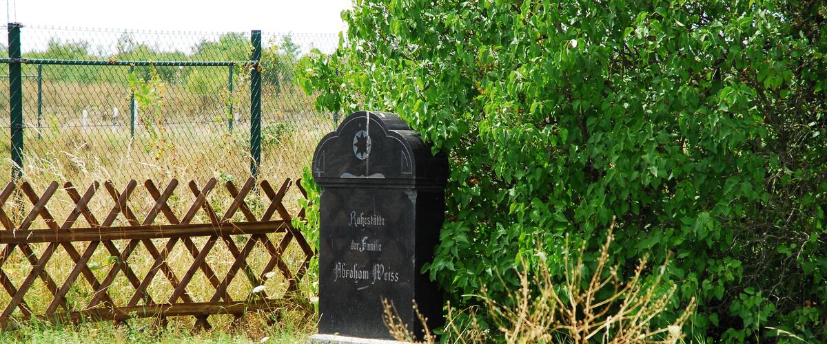 A headstone with inscriptions in a cemetery surrounded by greenery and a wooden fence.
