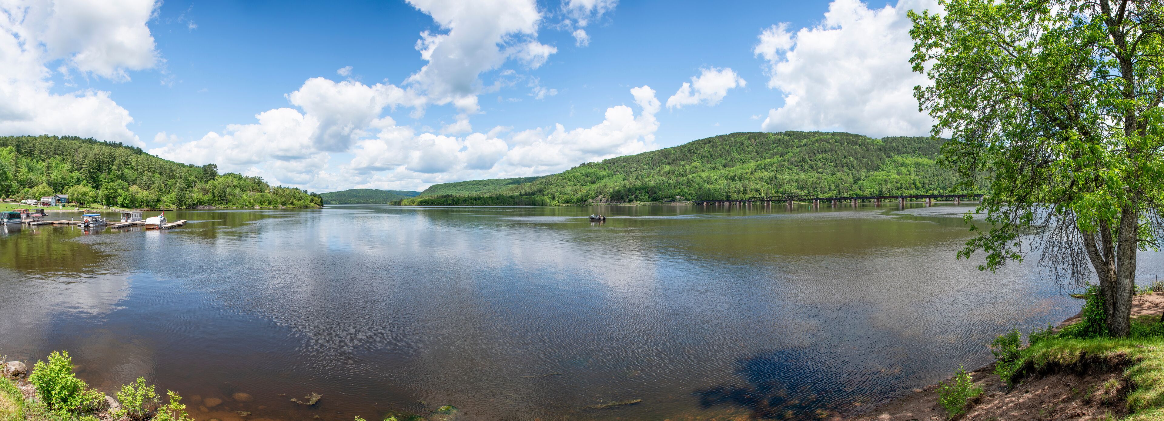 A calm lake with a wooden pier, surrounded by lush green hills and a clear blue sky.