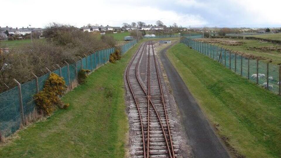 Railway tracks curving through a rural landscape with green fields and a fence along one side.