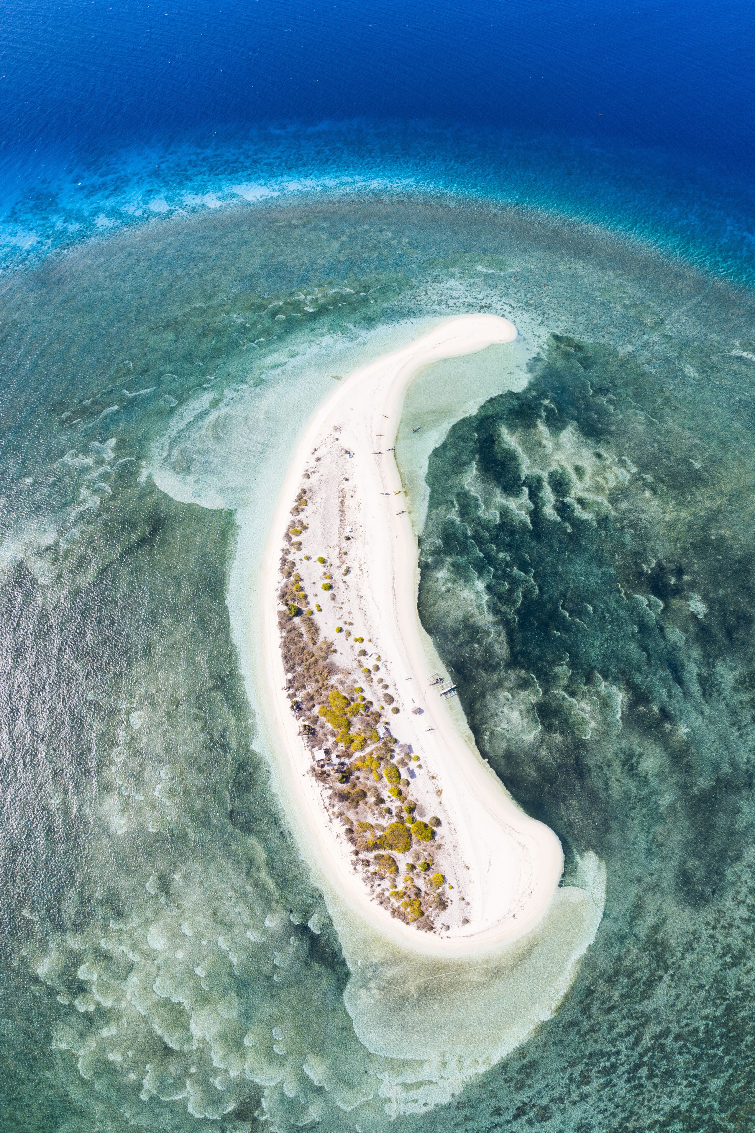 An aerial view of a narrow, white sandy beach curving into a turquoise sea, surrounded by clear shallow waters and a coral reef.