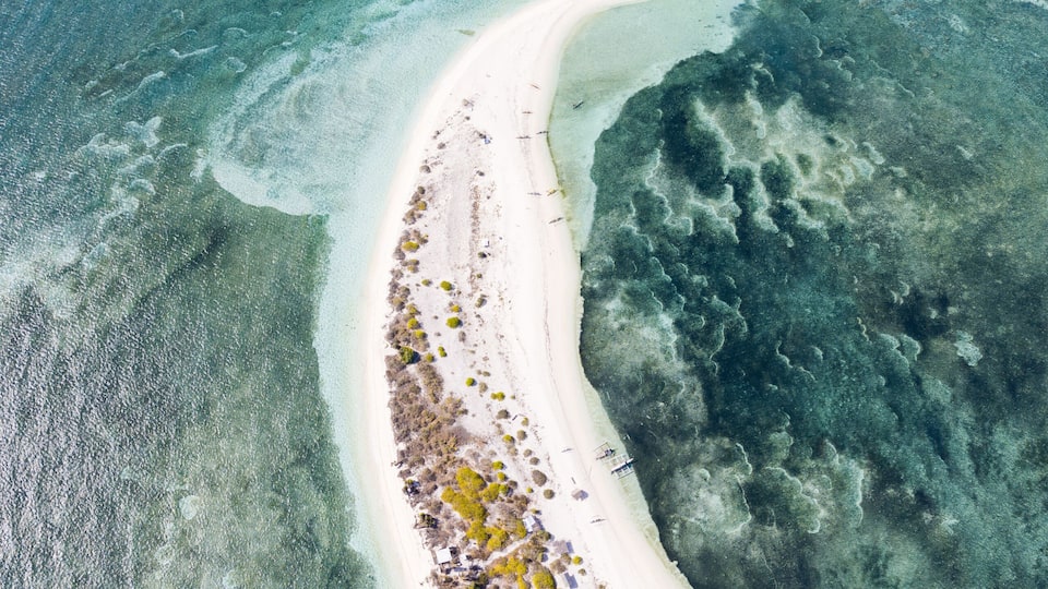 An aerial view of a narrow, white sandy beach curving into a turquoise sea, surrounded by clear shallow waters and a coral reef.