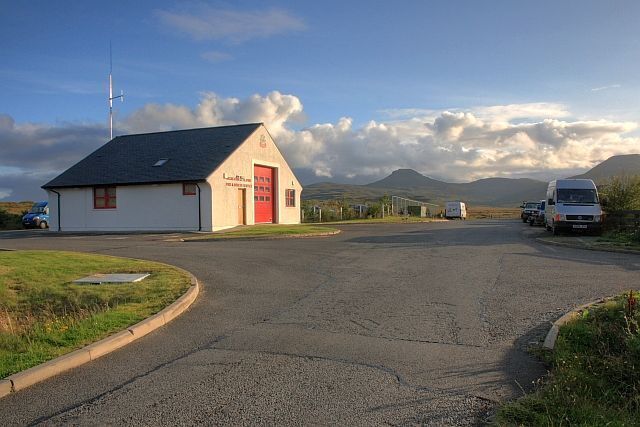 A rural road with a white building with a red door, a van, and a truck parked on the side.