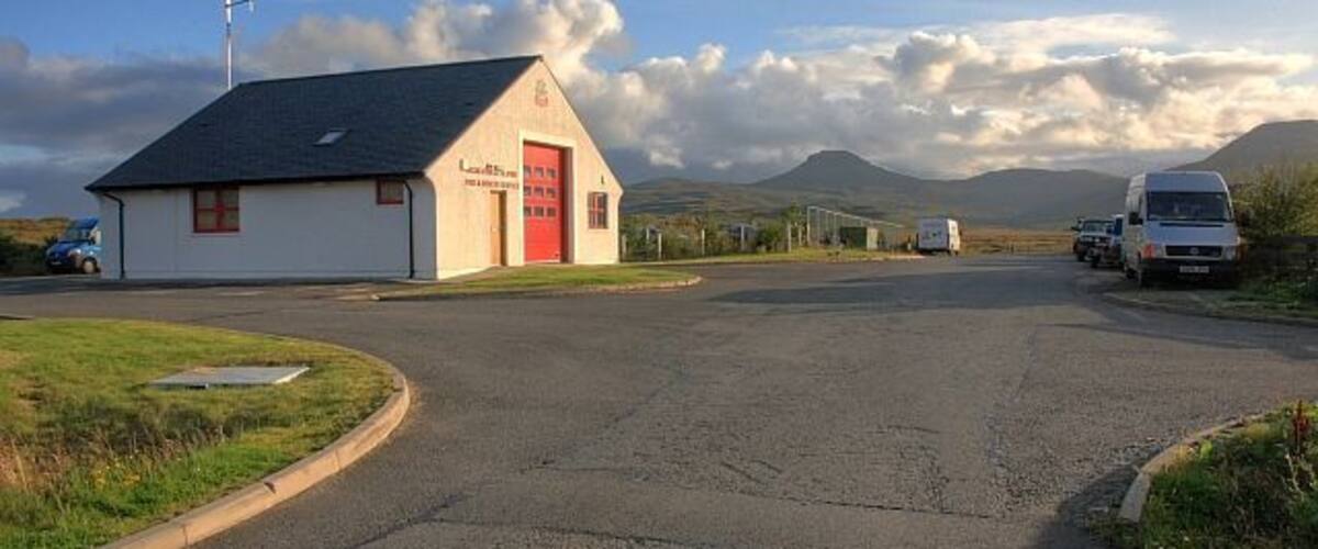A rural road with a white building with a red door, a van, and a truck parked on the side.
