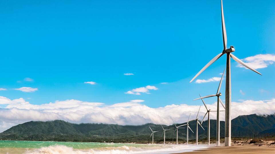 A beach with wind turbines and mountains in the background.